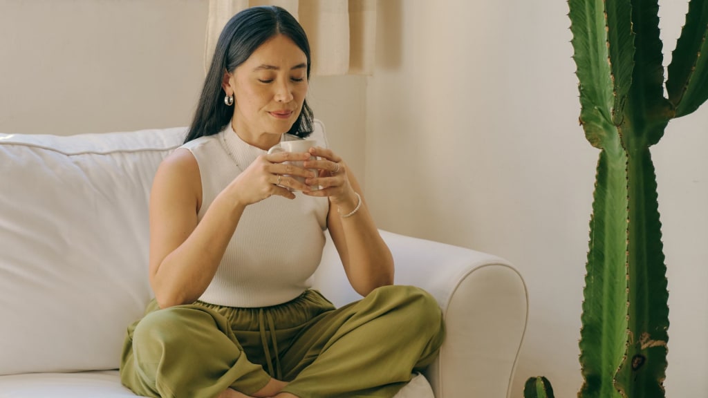Woman with long dark hair sits cross-legged on a white couch, holding a cup with both hands, next to a tall cactus in a brightly lit room—a serene setting perfect for a burnout therapist's calming retreat.